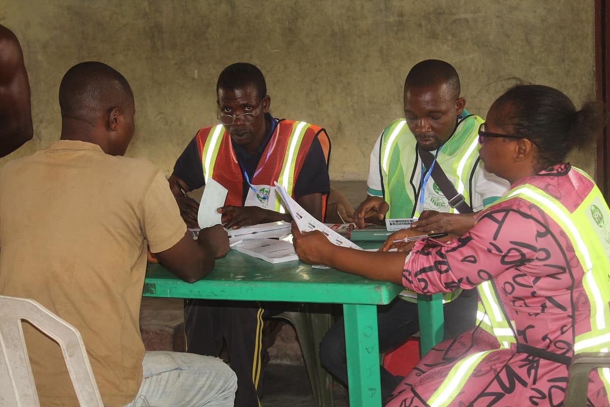 Polling officials in reflective INEC vests check a voter's details at a green table, holding the register and a voter's card.