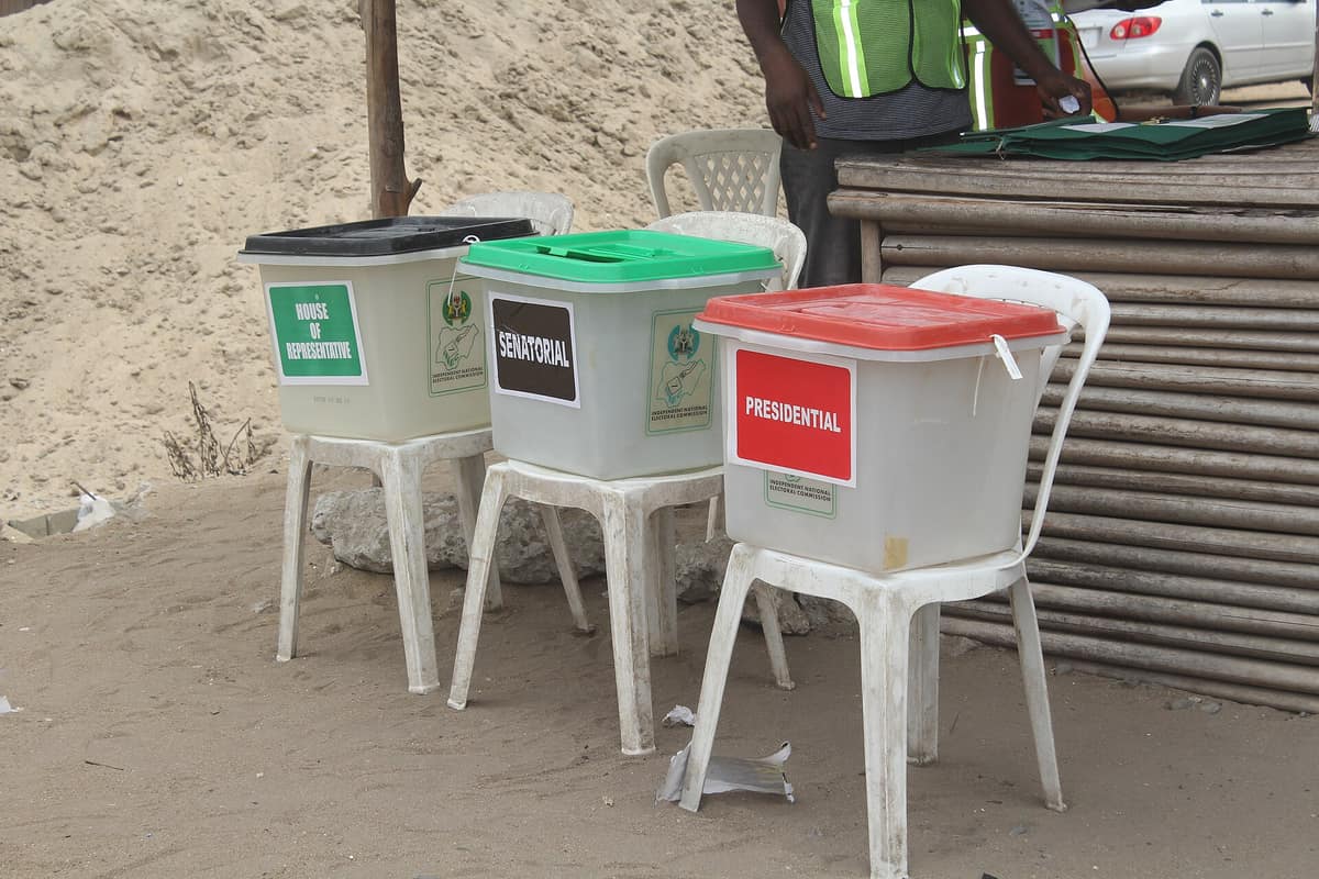 Three transparent INEC ballot boxes on white plastic chairs, labelled House of Representatives, Senatorial and Presidential.