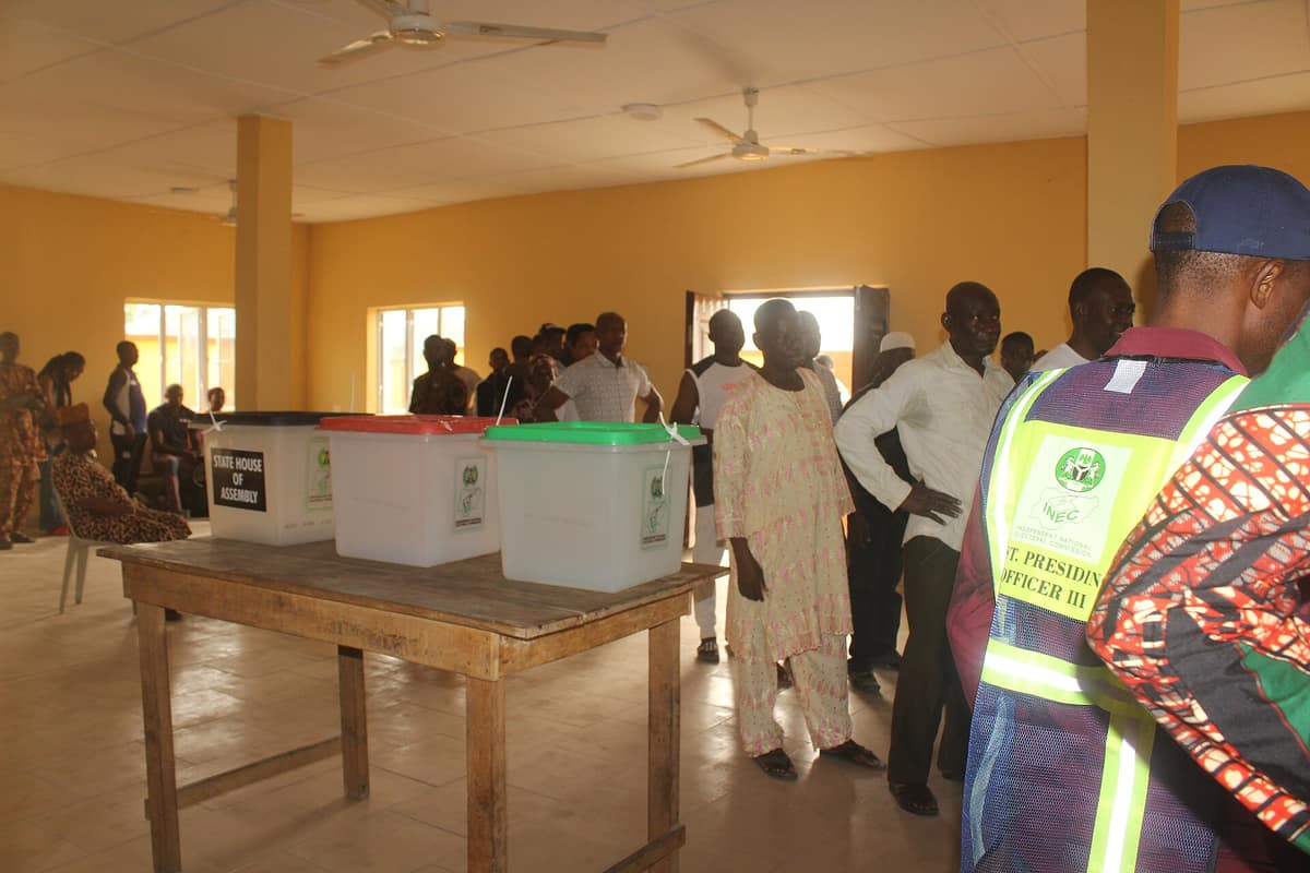 Voters stand in a yellow hall near ballot boxes on a wooden table while an assistant presiding officer in an INEC vest looks on.
