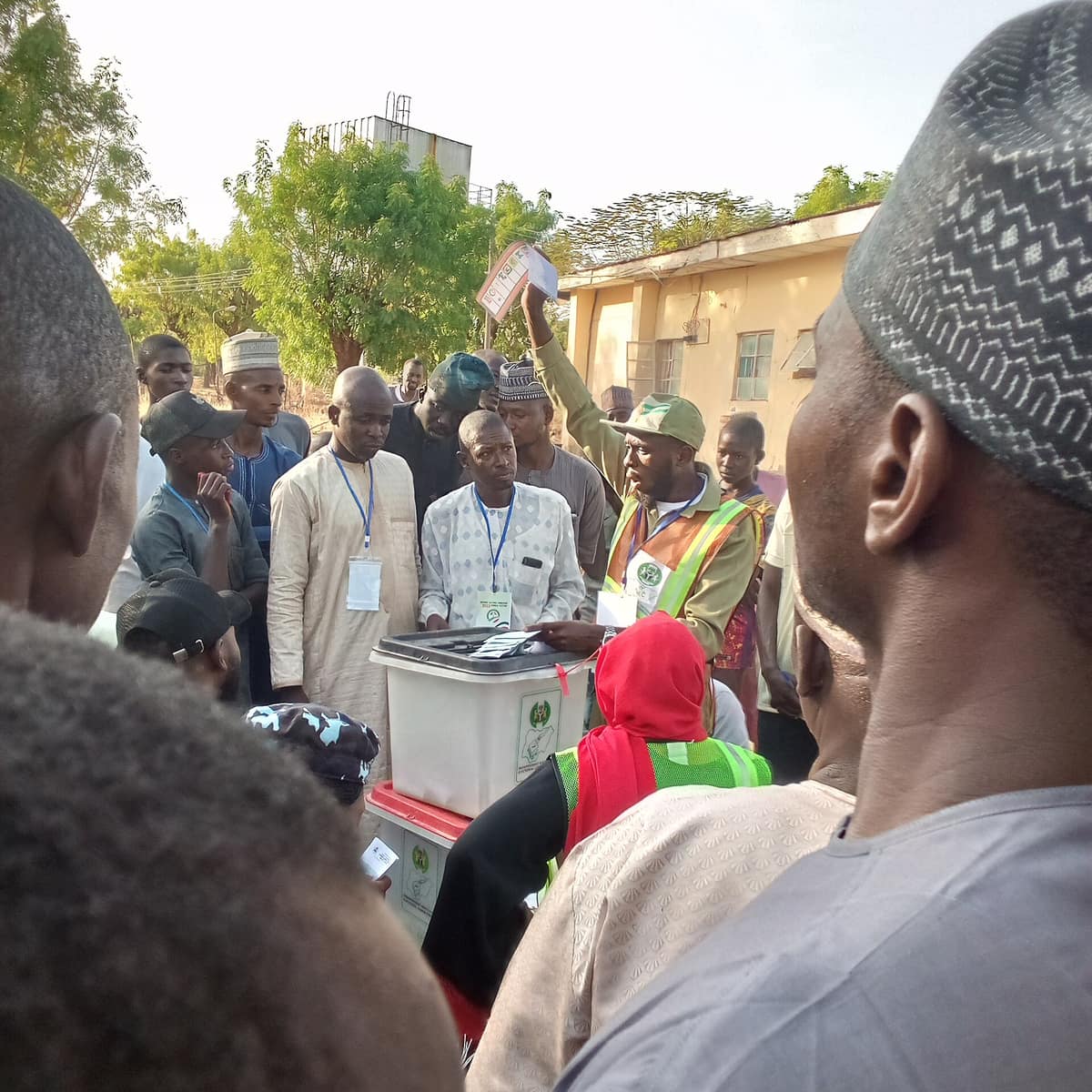 A polling official in an INEC vest holds a ballot paper high for all to see as agents and voters gather around the ballot box.