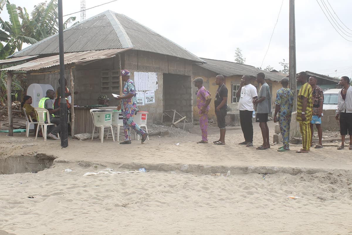 A line of voters waits on a sandy street beside a small polling unit with ballot boxes, officials at a table and a Polling Zone sign.