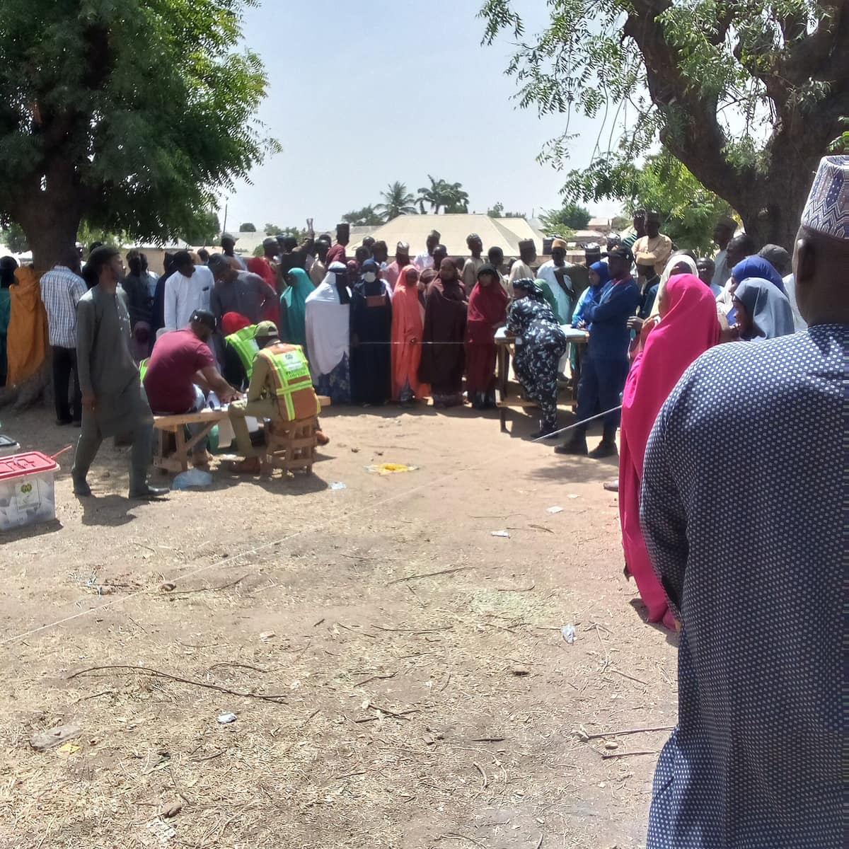 A large crowd of men and women, many in bright hijabs, waits behind a rope under shade trees while officials work at a bench.