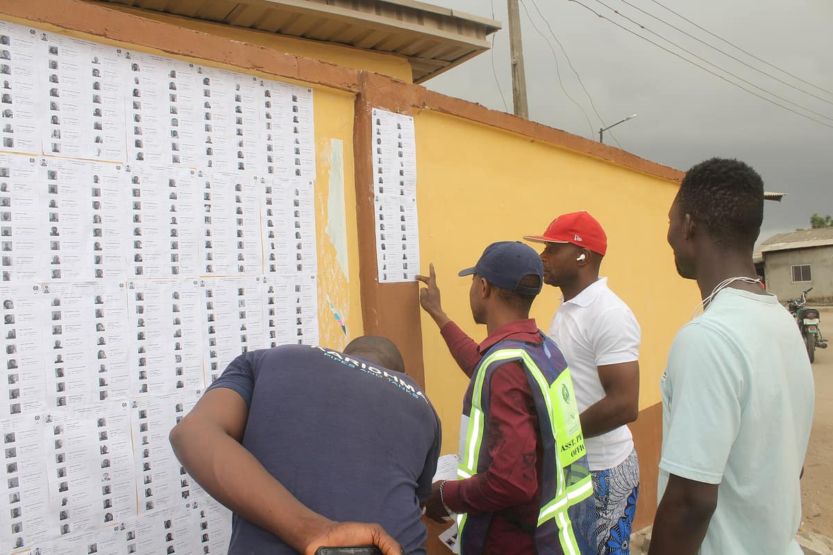 Voters and an assistant presiding officer search the printed voters' register pasted on a yellow wall outside a polling unit.