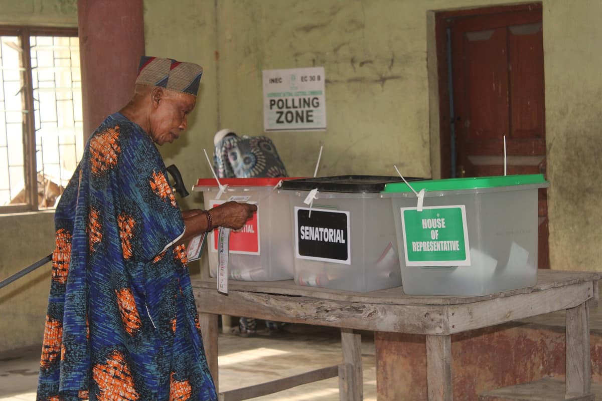 An elderly man in a blue and orange agbada drops his ballot into the Presidential box. Senatorial and House of Representatives boxes stand beside it under an INEC Polling Zone sign.